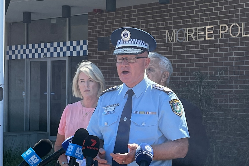 Police officer in hat speaks in front of bank of microphones