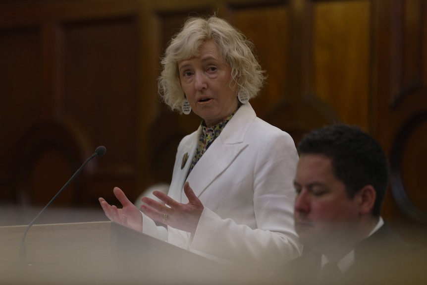 A blonde woman with a white jacket speaking at a lectern.