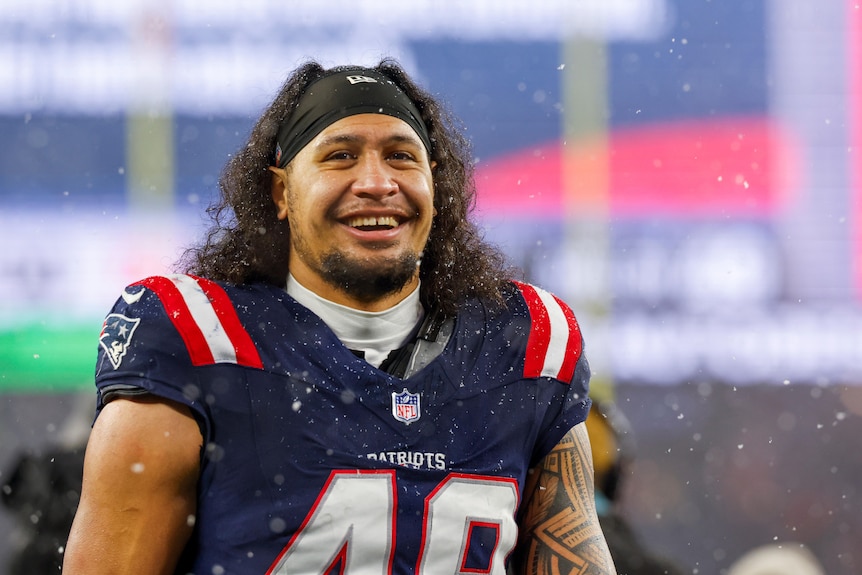 A Samoan man with long hair in a black hair band and wearing a Patriots NFL jersey smiles at the camera