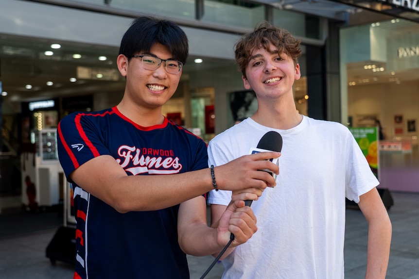 Two teenage boys, one with black hair and glasses, the other with brown hair and a white t-shrt, hold a mic and smile.