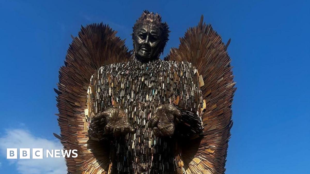 A tall male figure towers above a grey. The statues hands are open and shoulders are shrugged. The statue is made out of rusty knife blades of all different shapes and sizes.