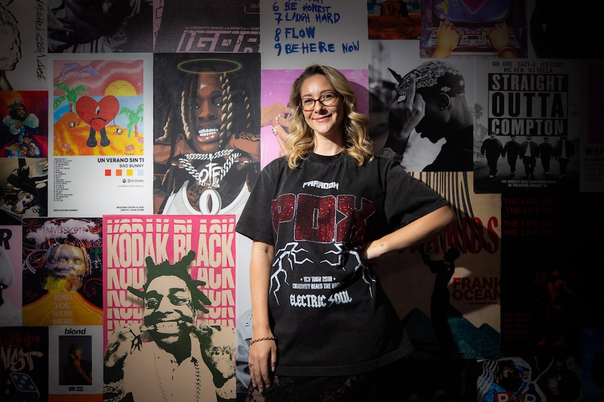 A woman in a black tshirt standing in front of wall with music posters on it