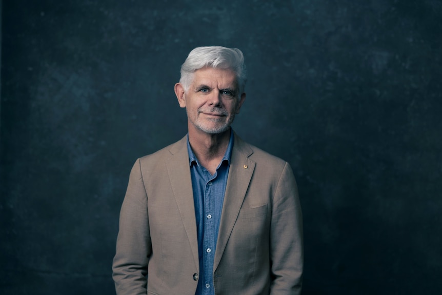 A man with silver hair, wearing a blue shirt and a tan blazer, standing in front of a dark blue background.