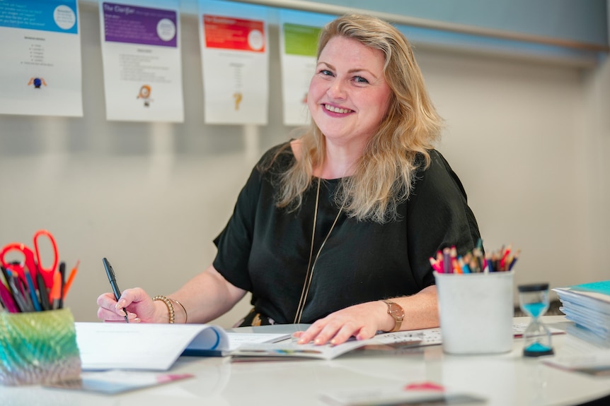 Eleanor in a dark top, sitting at a teacher's desk, smiling