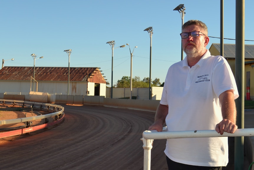 Regan wears a white shirt and glasses, standing beside the greyhound track, resting his hands on a railing.