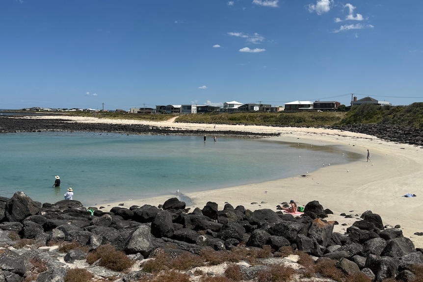 A sheltered beach, with clean sands surrounded by basalt rocks, clear blue sky, houses behind shurbs.
