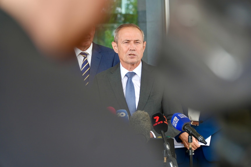 WA Premier Roger Cook speaking at a media conference with microphones in front of him and blurry people in the foreground.