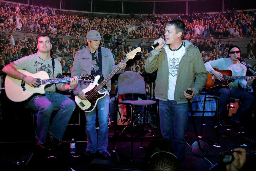 A grunge rock band in mid-2000s clothes performs in front of an auditorium of people