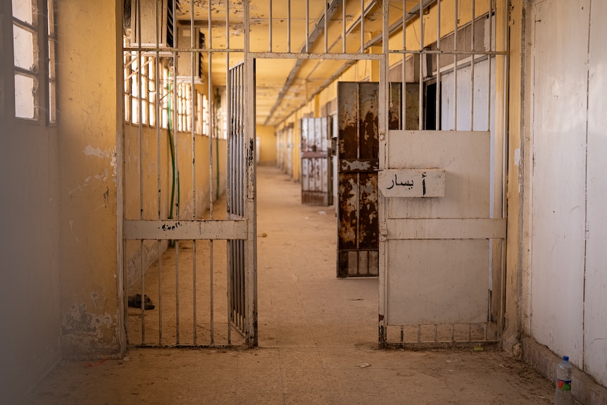 A prison hallway with unlocked gates and doors. The walls are yellow and there are windows to the left.