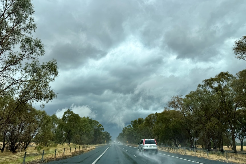 A car travels along a wet highway under stormy grey clouds hanging low over trees.