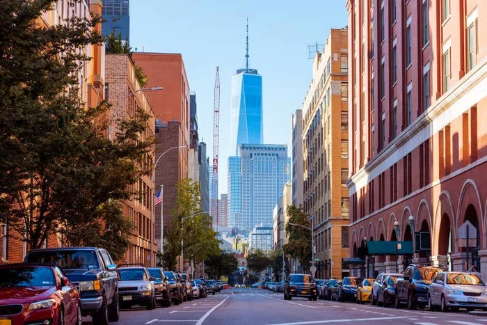 Street view with One World Trade Center towering in the distance, framed by urban buildings and parked cars