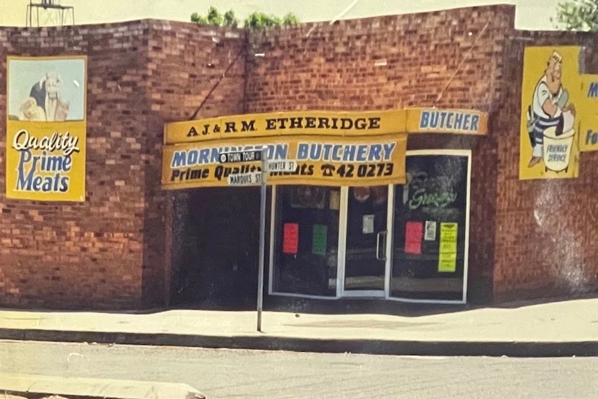 old photo of red brick Mornington Butchery shopfront. Shop is decorated with colourful signage and cartoon butcher character.