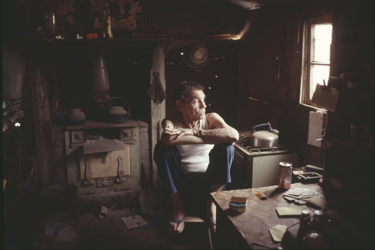 A man in a singlet sits in his home and looks out the window. 