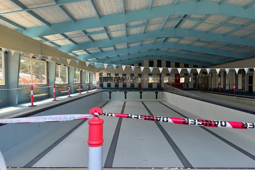 An empty swimming pool surrounded by "danger" tape, cream and turquoise rafters.