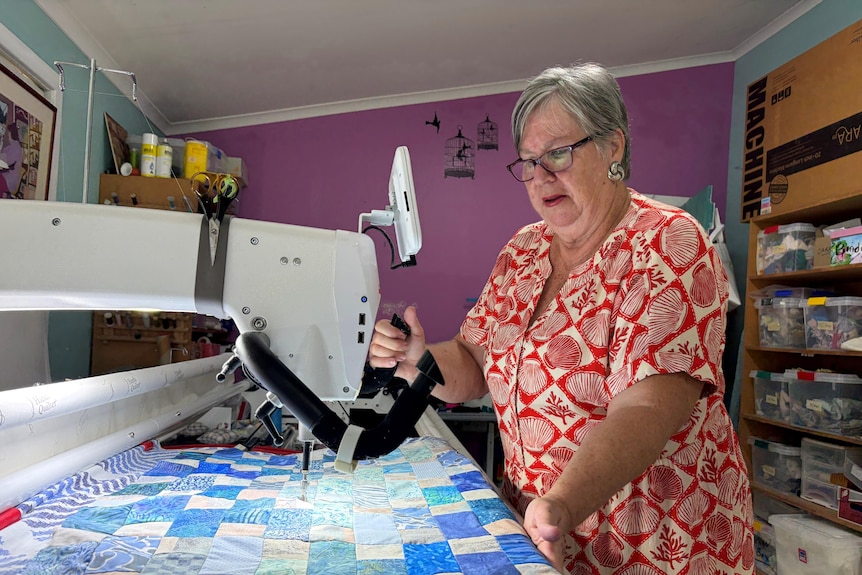 An older woman standing at a quilting machine, with a bright coloured quilt.