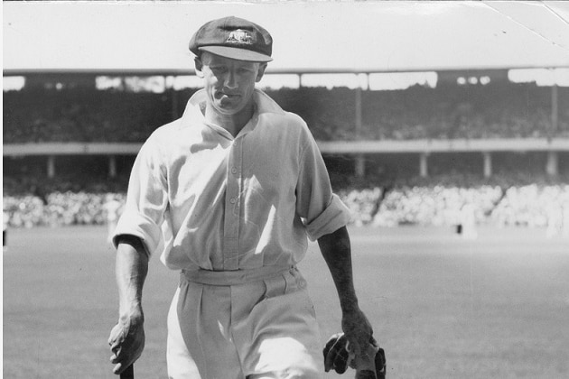 Don Bradman walking off with bat and gloves in his hands, baggy green on his head.
