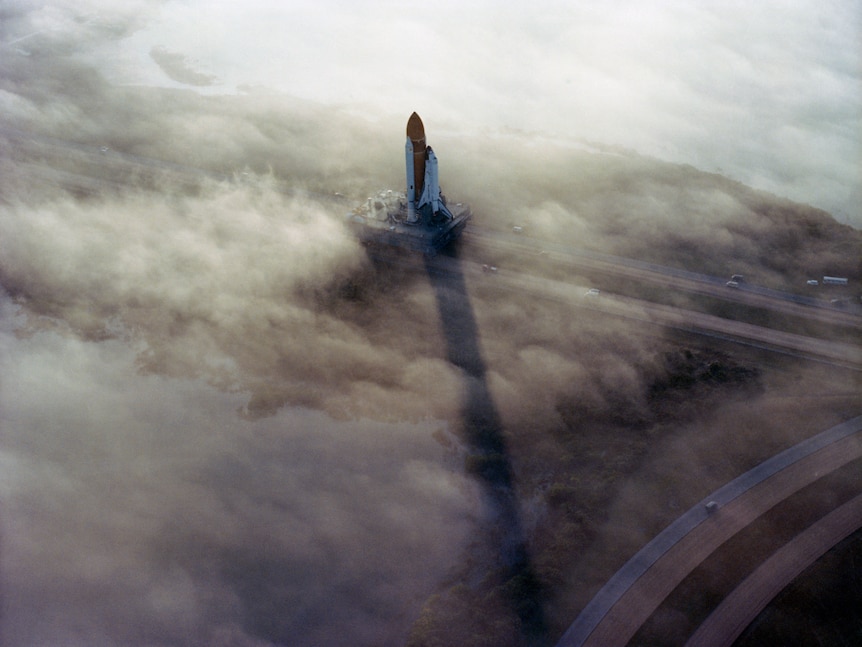 In an aerial photo, a space shuttle is seen in the distance looming vertically out of fog and casting a long shadow