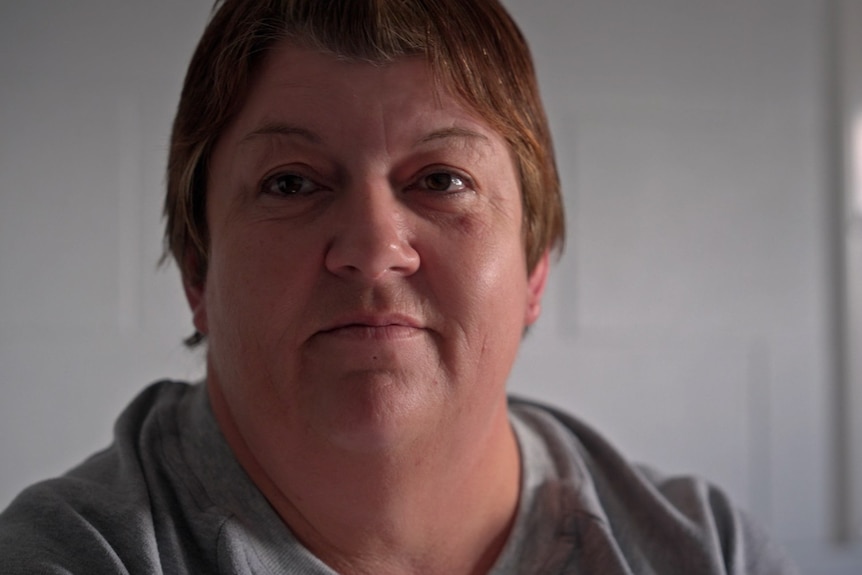 A short haired woman looks slightly to the left while wearing a grey top and standing in front of a whiteboard, misty eyed.