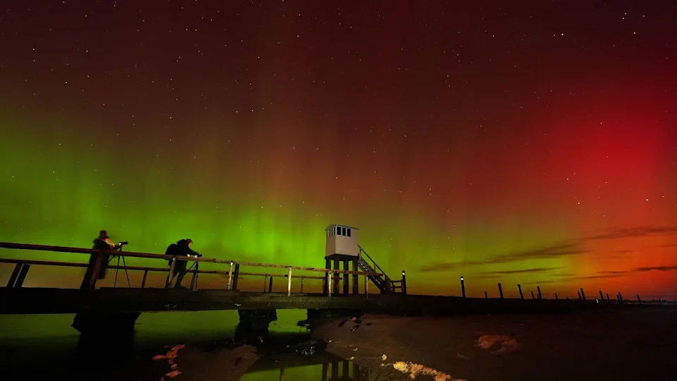 Two people taking photographs near a white hut, raised on stilts. The sky is lit red and green.