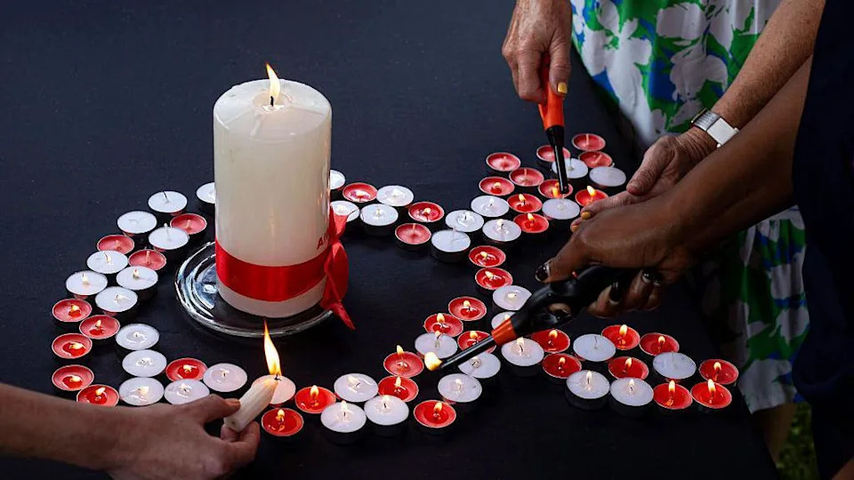Small red and white candles are arranged in the shape of an Aids ribbon. A large white candle is in the centre. The candles are being lit by some disembodied hands. 