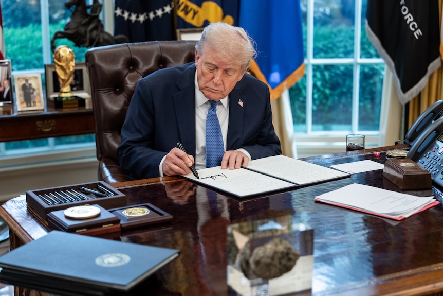 Donald Trump signs a document in the oval office. On his desk sits a rock-shaped lump encased in a glass-like cube.