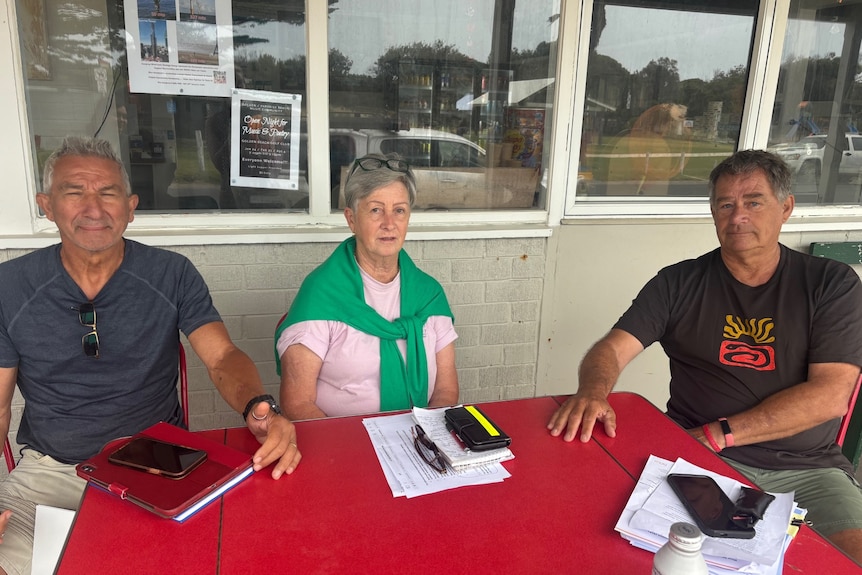 Two men and a woman sit around a table, outside a local corner store. 