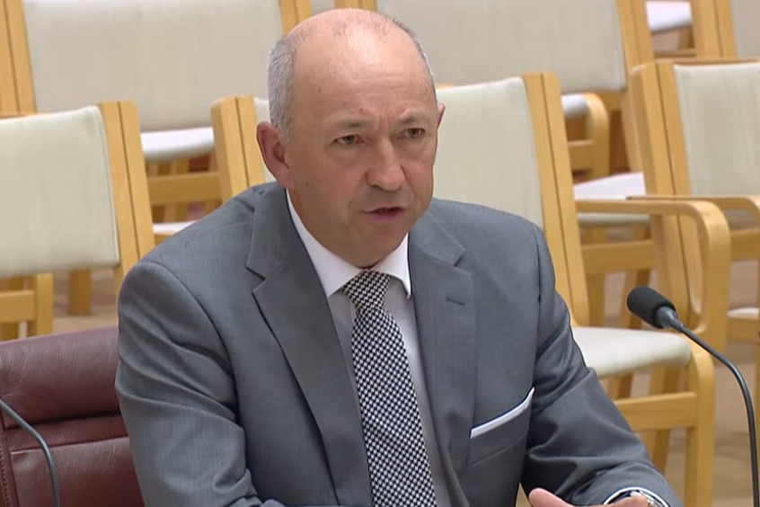 A balding man in a grey suit talks into a microphone