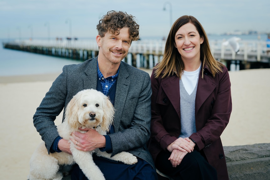 Leon, left, holds a white doodle with Celia, right, while the pair sit on a stone wall at a beach with a jetty behind them.