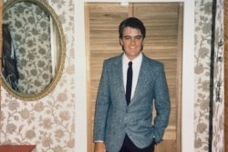 a young man in a grey suit poses in a bedroom