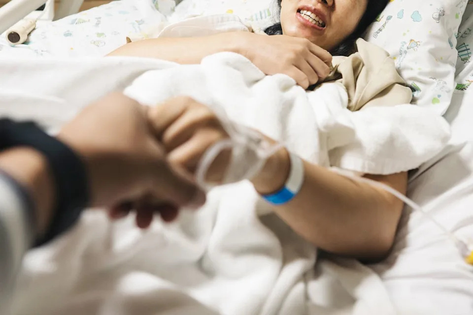 A woman in pain in a hospital bed holding the hand of another person (stock image). Getty