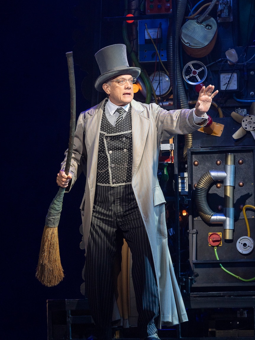 On stage, Simon Burke, early 60s, in a top hat, glasses and coat, holds out one arm, the other holding a broomstick.