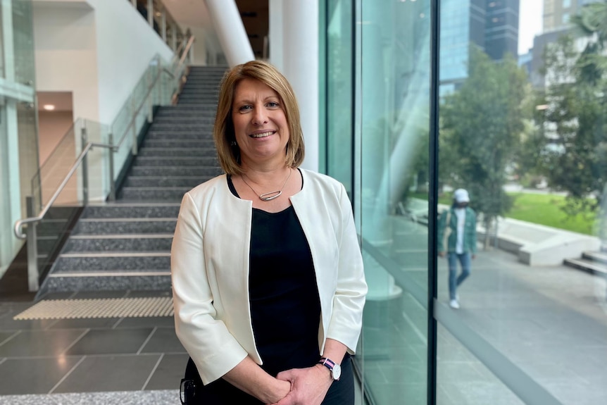 A woman wearing a black dress and a cream jacket stands inside a glass office building.
