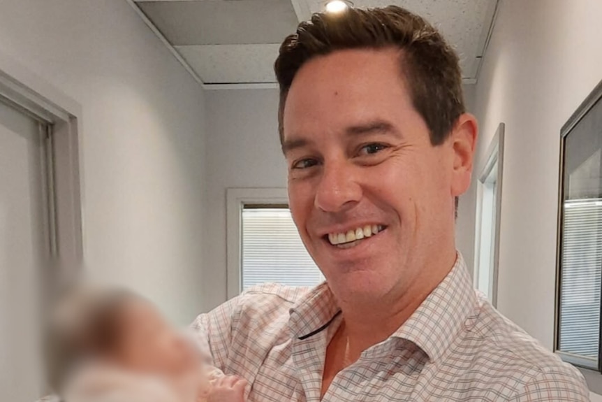 A man wearing a collared shirt smiles for the camera holding a blurred baby.