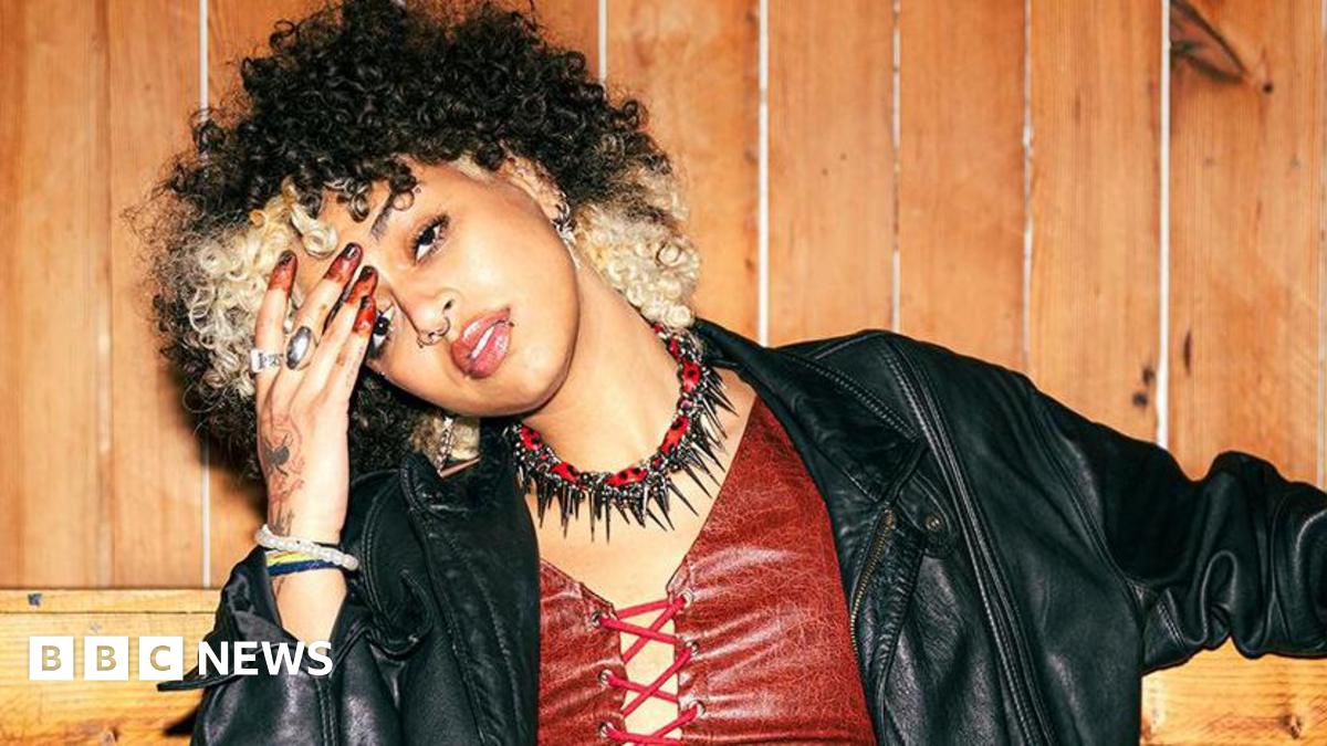 Nxdia with blond and black hair rests her head to the side on her hand. She is wearing a red leather-look top with a leather jacket and black and red choker necklace with spikes.