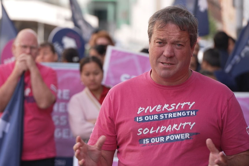 A man with short grey and brown hair stands in front of a rally of protestors in a pink HACSU shirt.