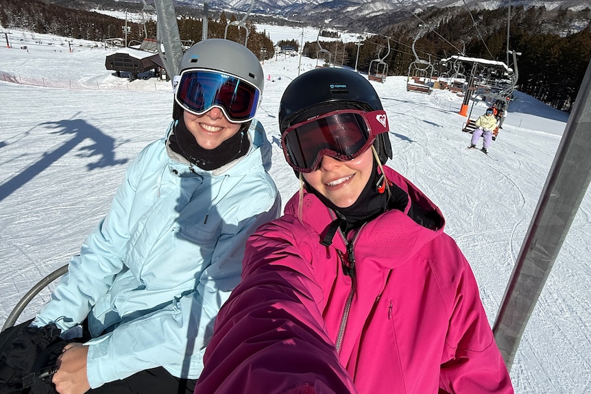 Two young women in ski clothes sit on a ski lift surrounded by snow.