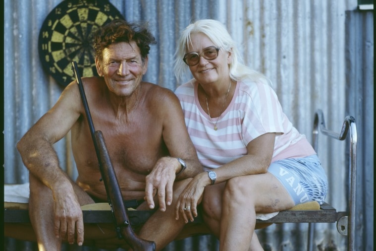 A couple sit outside a tin shed and smile at the camera, a shot gun leaning on the man.