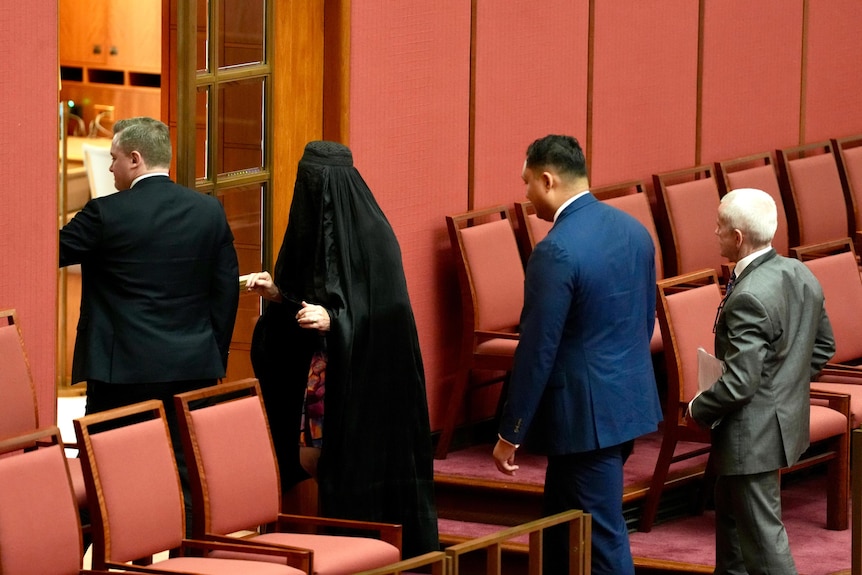 A woman in a dark burka — Pauline Hanson — files out of a parliament chamber along with several men in suits.