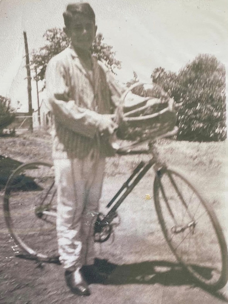 Black and white photograph of boy standing beside a bicycle with a basket. He is dressed in striped pants and a striped jacket.