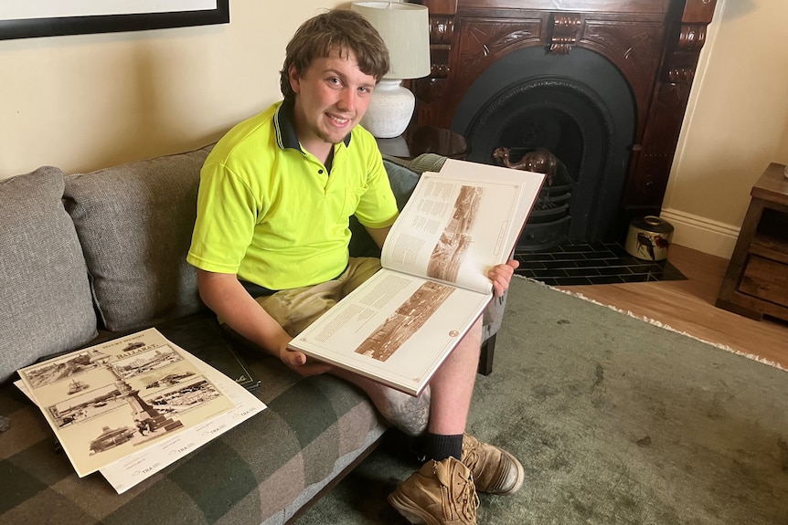 Young man in workwear sitting on a couch, scanning a history book. 