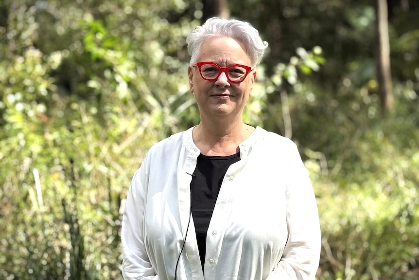 Woman wearing red glasses and grey hair in bushy area.