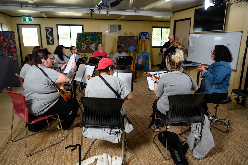 A group of women sat in a circle playing ukuleles and looking at sheet music with a man in the back corner playing a violin.