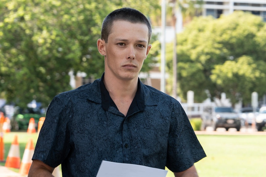 A young man in a button-up shirt, walking out of court while holding a piece of paper.