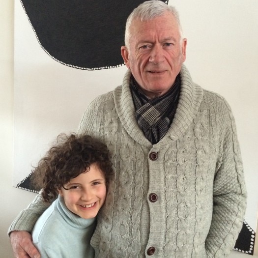 A grandfather wearing a grey knit jumper and scarf stands next to their smiling grandchild