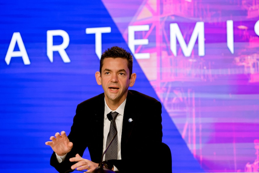 A man in black suit gestures with his hands as he sits and speaks in front of the Artemis logo.