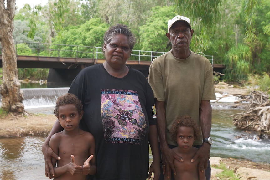 A couple with two young boys look seriously at the camera from in front of a bridge.