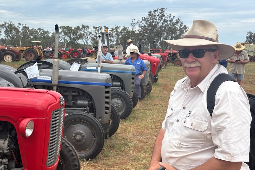 Peter stands in-front of a line of old tractors wearing a big hat, backpack and sunglasses. 