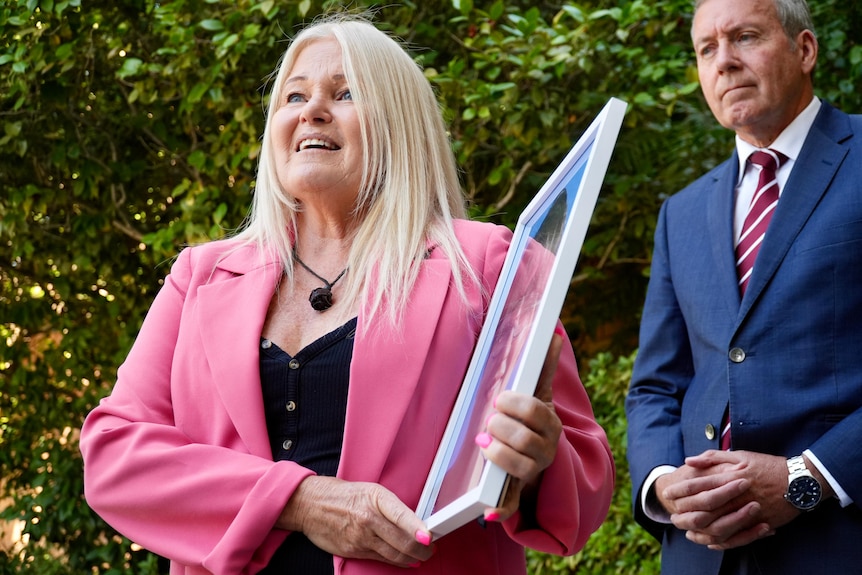 A woman with blonde hair, a pink jacket and a black shirt, holding a picture frame, stands talking in front of Reece Whitby.