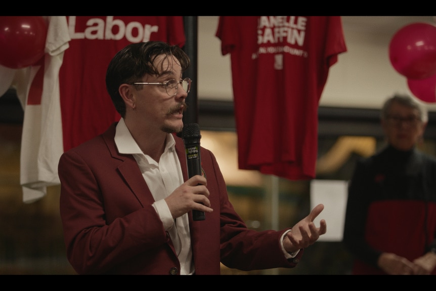 A film still of Harper Dalton, in glasses and a maroon blazer, speaking into a microphone at a community hall.
