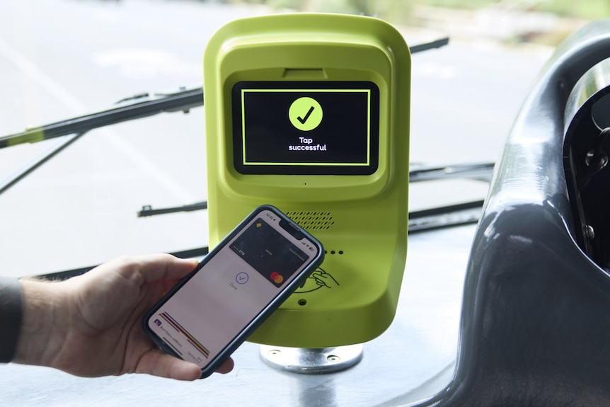 A hand holds up a mobile phone which is displaying a credit card to a yellow payment screen on a bus that has a tick on it.